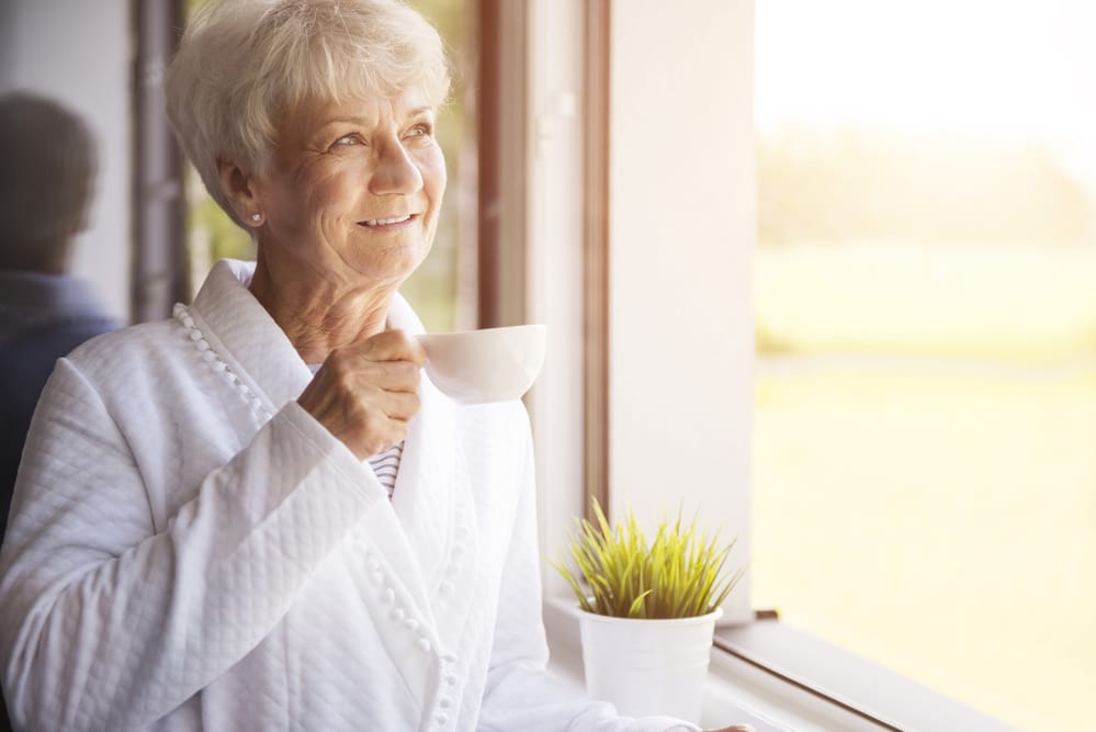 Smiling senior woman in robe holding coffee looking out window in the morning