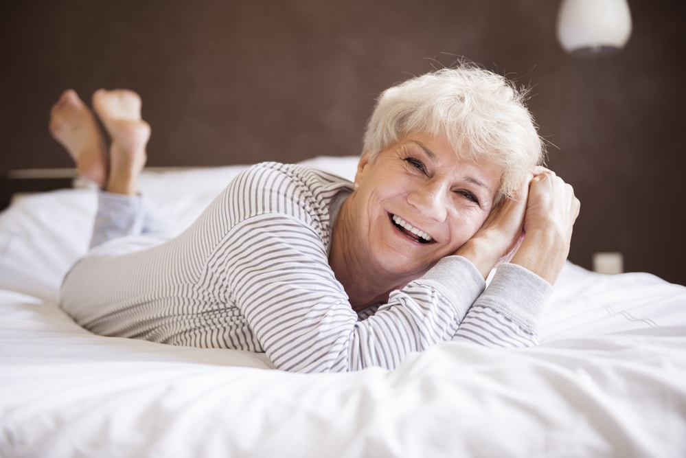 Senior woman smiling, lying on stomach in bed, wearing pajamas