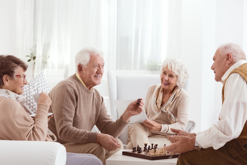 Four smiling seniors, two men playing chess and two women watching